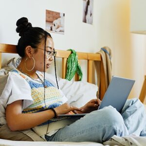 Photograph of a girl sitting on a bed, typing on a laptop computer.