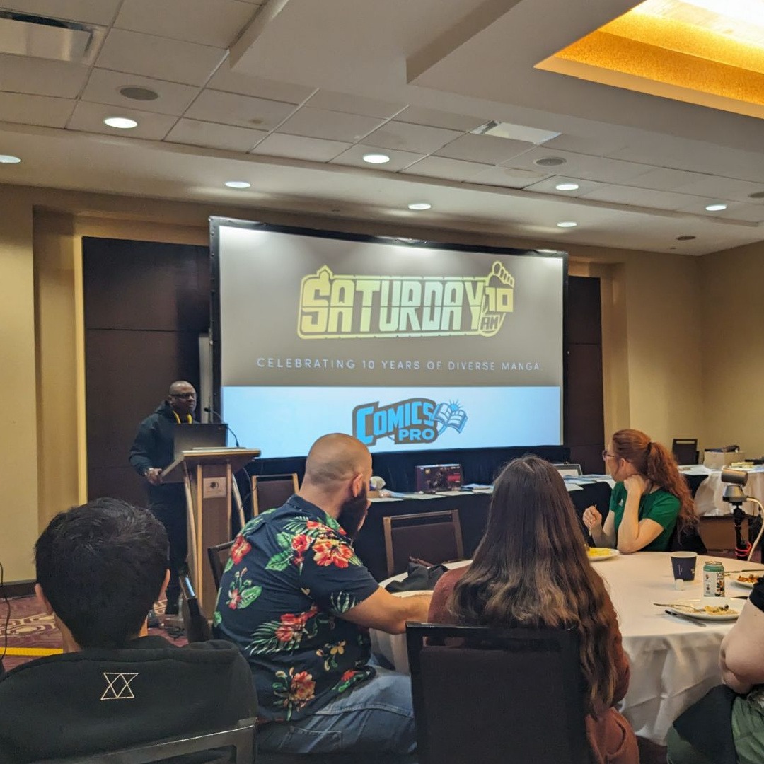 Photo of Frederick L. Jones standing alongside a screen with a projected image of the Saturday AM logo, as he makes a presentation at the ComicsPRO conference.