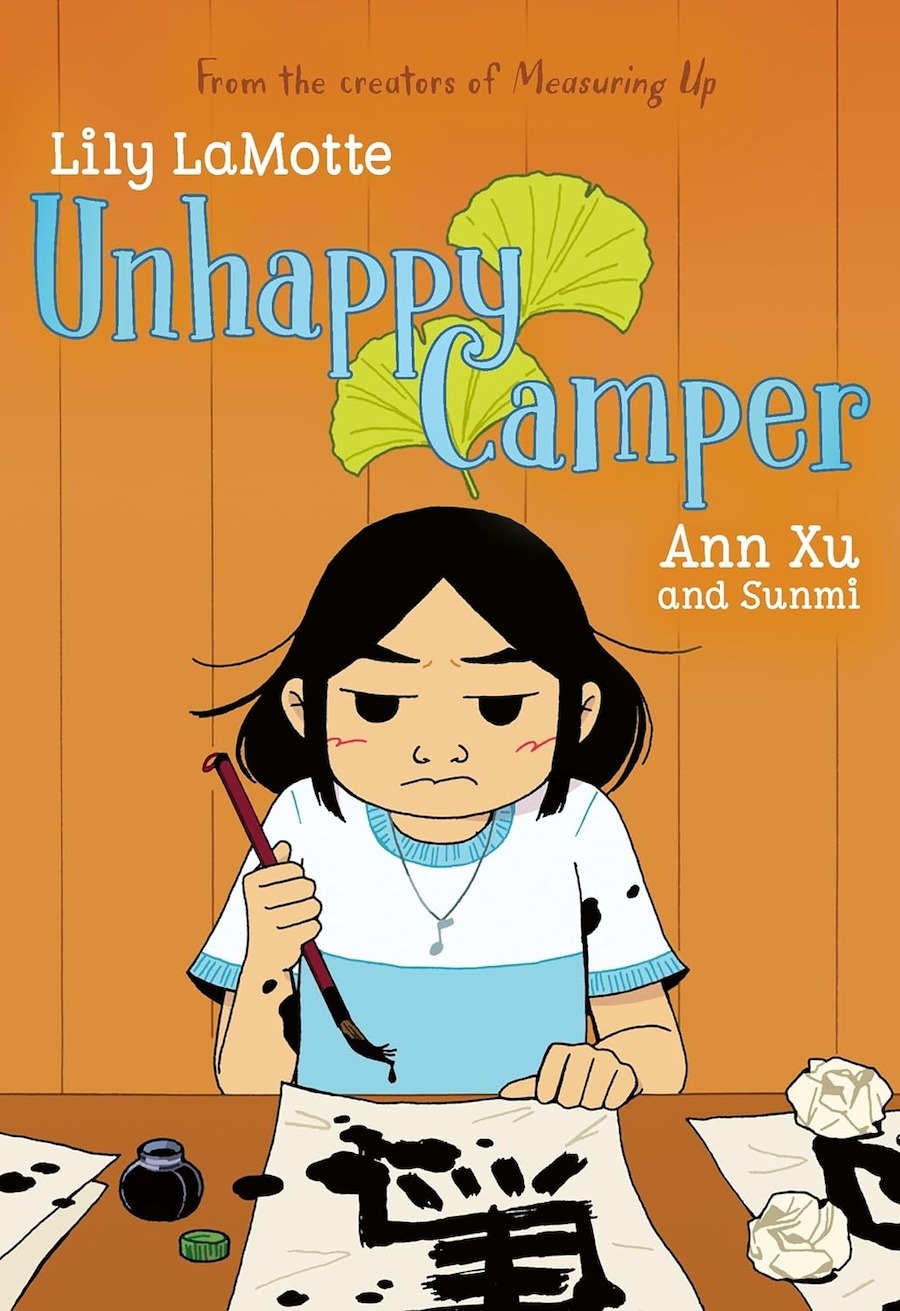 Cover of Unhappy Camper, showing an Asian Girl frowning as she holds a brush over a sheet of calligraphy; the brush is dripping and there are spots of ink on the paper where she has been drawing the characters.